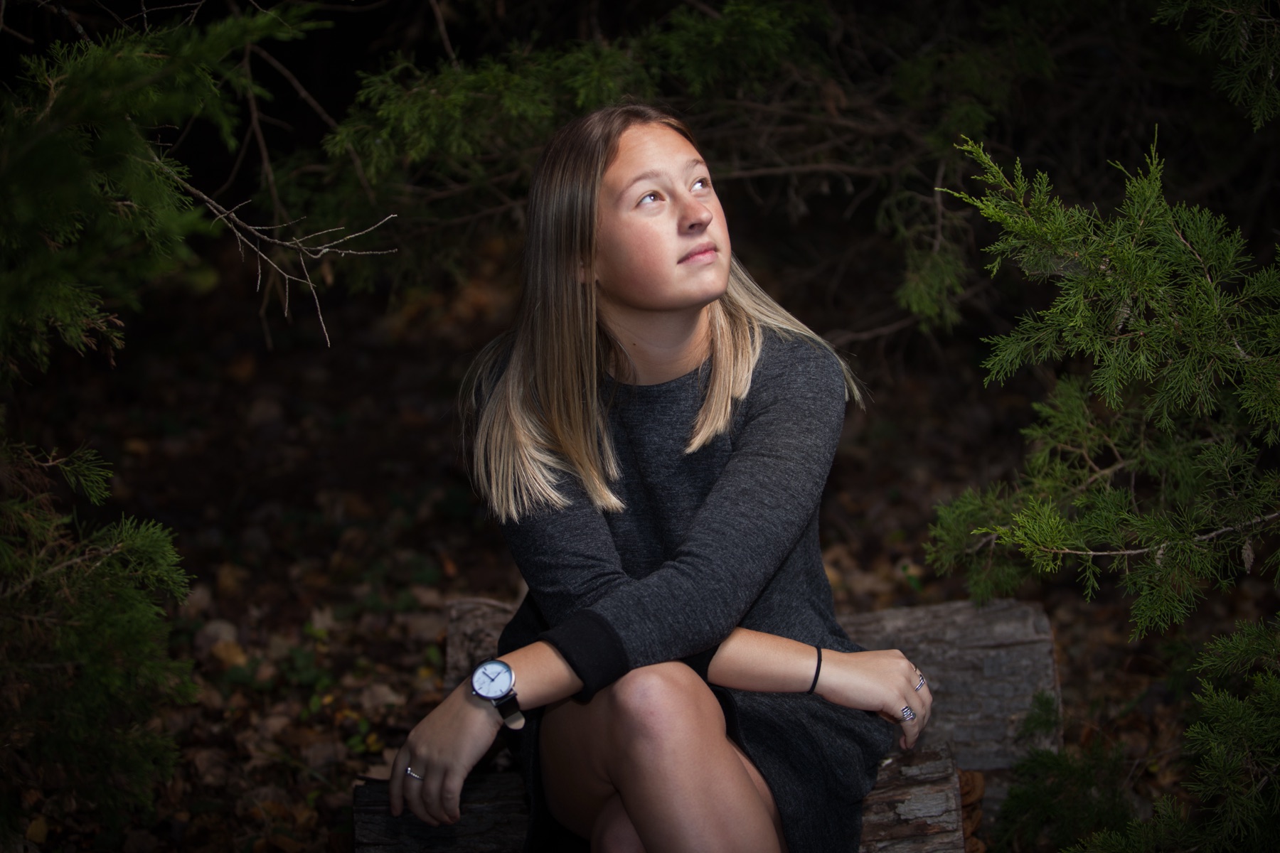 Young woman in a gray sweater dress sitting in a shaded wood, looking up toward the light