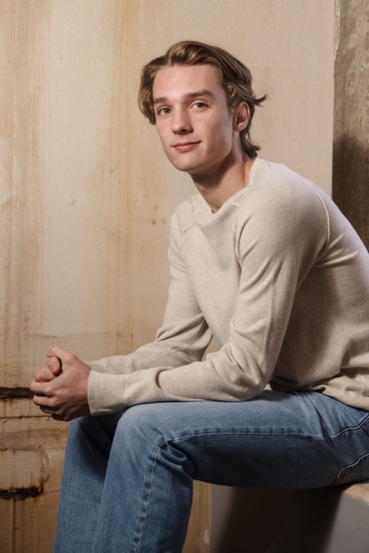 Young man in a cream sweater and jeans sitting against a weathered wall