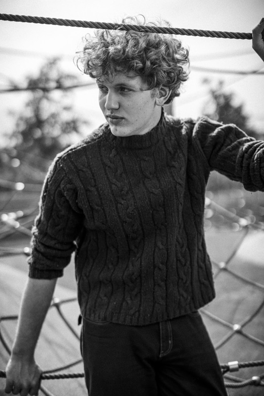 Black-and-white portrait of a young man with curly hair in a cable-knit sweater on a rope net