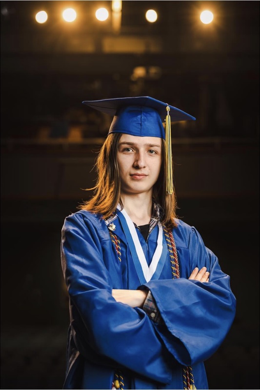 Graduate in a blue cap and gown with honor cords, arms crossed, in a dim theater