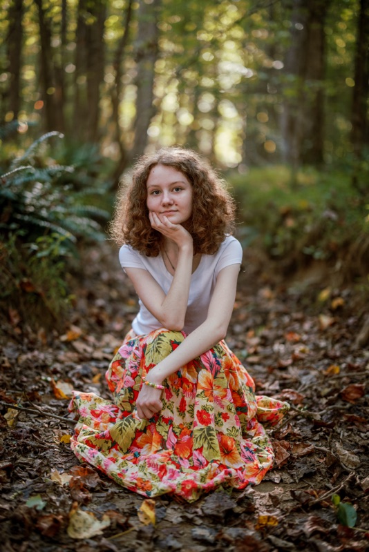 Young woman in a floral skirt sitting among fallen leaves with her chin in her hand