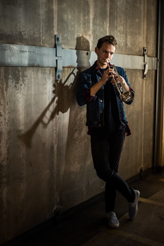Young man playing trumpet against a concrete wall, his shadow beside him