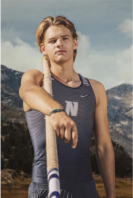 Pole vaulter in a striped team singlet resting his pole on his shoulder in front of mountains
