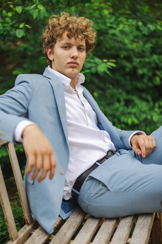 Young man in a light blue suit lounging on a wooden bench in front of greenery