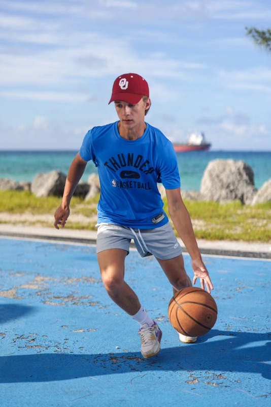 Young man in a red cap and blue T-shirt dribbling a basketball on an outdoor court by the sea